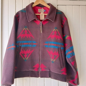 Puede incluir: Chaqueta de lana granate con un estampado geom&eacute;trico en rojo, turquesa y azul. La chaqueta tiene cremallera frontal, cuello y dos bolsillos. La chaqueta est&aacute; colgada de una percha de madera.