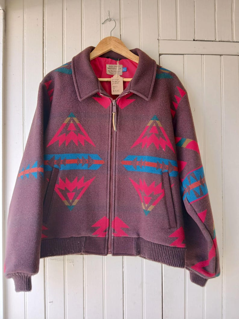 Puede incluir: Chaqueta de lana granate con un estampado geom&eacute;trico en rojo, turquesa y azul. La chaqueta tiene cremallera frontal, cuello y dos bolsillos. La chaqueta est&aacute; colgada de una percha de madera.