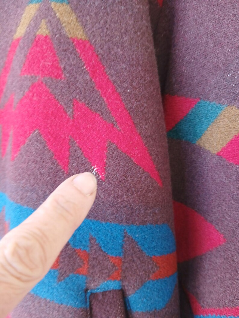 Puede incluir: Primer plano de una secci&oacute;n de tela estampada. La tela es de color p&uacute;rpura oscuro con un dise&ntilde;o geom&eacute;trico en tonos rosa, turquesa y beige. El dise&ntilde;o incluye zigzags y formas de estrella. La tela parece ser un material tejido.