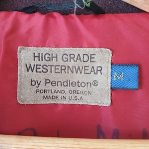 Puede incluir: Primer plano de una etiqueta de una chaqueta Westernwear Pendleton. La etiqueta es de color beige con texto negro: "HIGH GRADE WESTERNWEAR by Pendleton&reg; PORTLAND, OREGON MADE IN U.S.A." Una etiqueta azul con una "M" blanca est&aacute; adjunta.