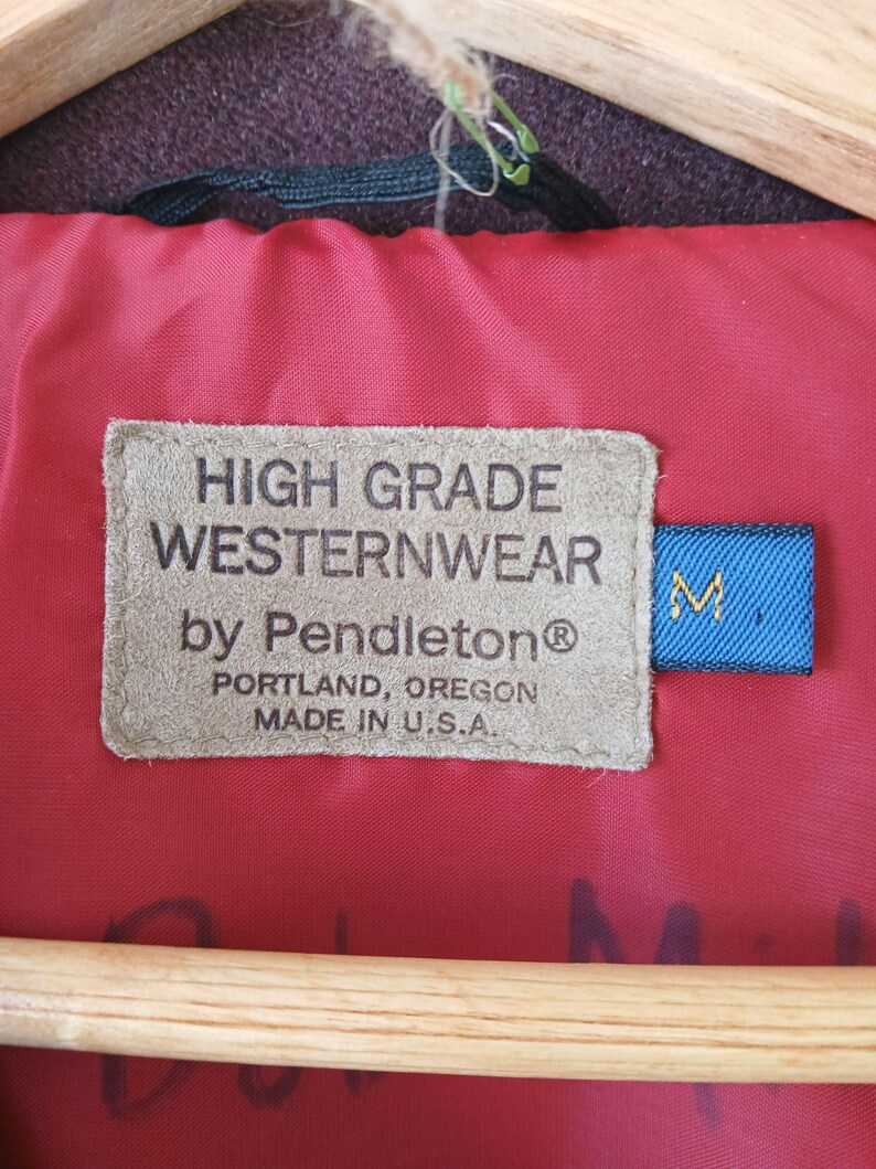 Puede incluir: Primer plano de una etiqueta de una chaqueta Westernwear Pendleton. La etiqueta es de color beige con texto negro: "HIGH GRADE WESTERNWEAR by Pendleton&reg; PORTLAND, OREGON MADE IN U.S.A." Una etiqueta azul con una "M" blanca est&aacute; adjunta.