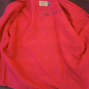 Puede incluir: Vista interior de un abrigo de lana marr&oacute;n con un forro rojo brillante. Una etiqueta dice "HIGH GRADE WESTERNWEAR" y "Bob Miller" est&aacute; escrito a mano en el forro. El abrigo est&aacute; colgado sobre una superficie de madera.