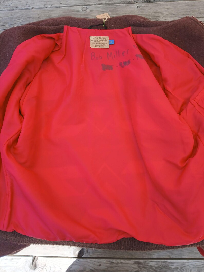 Puede incluir: Vista interior de un abrigo de lana marr&oacute;n con un forro rojo brillante. Una etiqueta dice "HIGH GRADE WESTERNWEAR" y "Bob Miller" est&aacute; escrito a mano en el forro. El abrigo est&aacute; colgado sobre una superficie de madera.