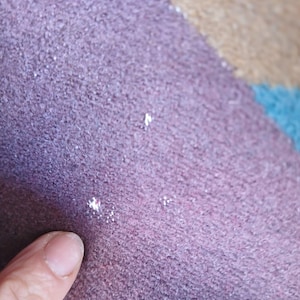 Puede incluir: Primer plano de una secci&oacute;n de una alfombra multicolor. La alfombra tiene una superficie texturizada con una paleta de colores p&uacute;rpura, rosa, beige y azul. La textura parece ser un material tejido o de punto. Un dedo se&ntilde;ala la alfombra.