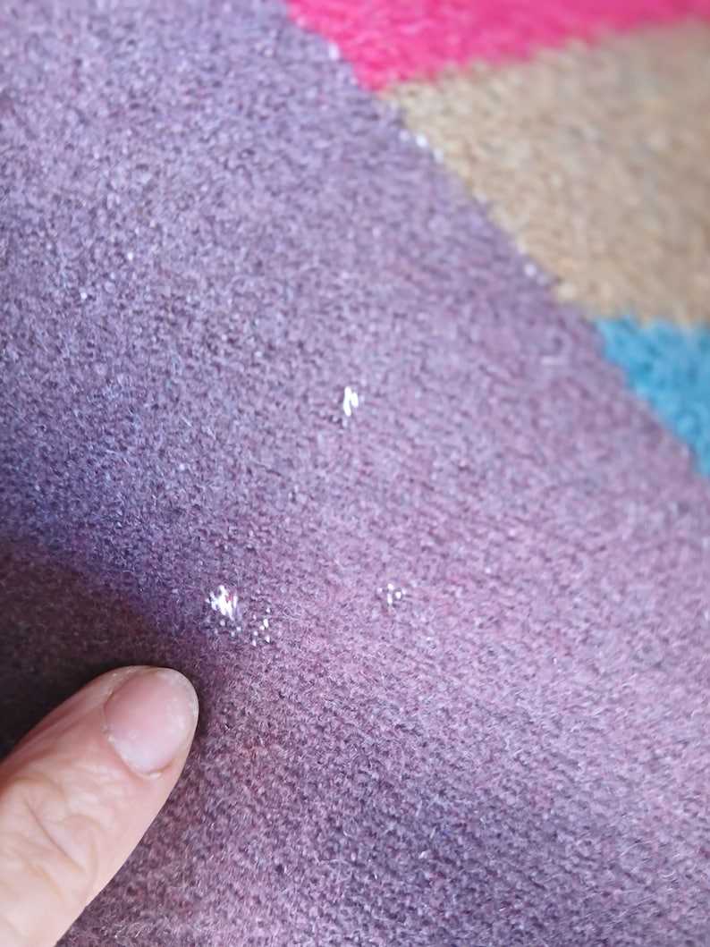 Puede incluir: Primer plano de una secci&oacute;n de una alfombra multicolor. La alfombra tiene una superficie texturizada con una paleta de colores p&uacute;rpura, rosa, beige y azul. La textura parece ser un material tejido o de punto. Un dedo se&ntilde;ala la alfombra.