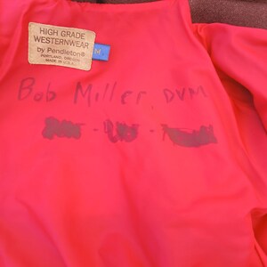 Puede incluir: Chaqueta de lana color burdeos con forro rosa brillante. El forro presenta una etiqueta que dice "HIGH GRADE WESTERNWEAR by Pendleton&reg;" y el nombre "Bob Miller, DVM" est&aacute; escrito a mano en el forro. Tambi&eacute;n es visible una etiqueta azul con una "M".