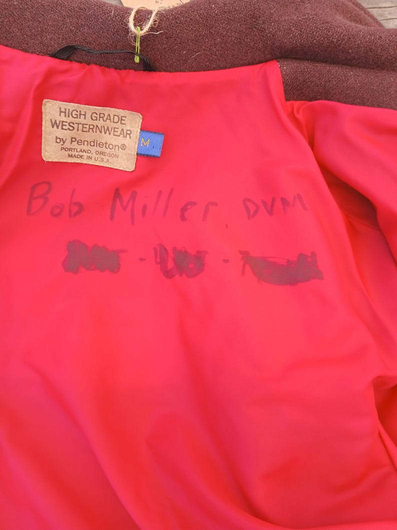 Puede incluir: Chaqueta de lana color burdeos con forro rosa brillante. El forro presenta una etiqueta que dice "HIGH GRADE WESTERNWEAR by Pendleton&reg;" y el nombre "Bob Miller, DVM" est&aacute; escrito a mano en el forro. Tambi&eacute;n es visible una etiqueta azul con una "M".