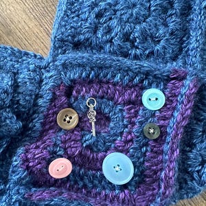 Op de afbeelding: Close-up van een handgemaakt gehaakt item in blauw en paars. Het item heeft een vierkante zak met een paars midden en diverse gekleurde knopen. Een zilveren sleutelbedel en een vlinderbedel zijn ook zichtbaar.