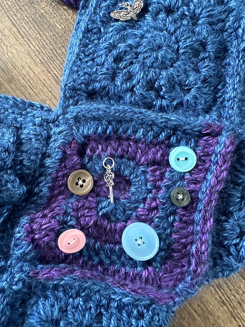 Op de afbeelding: Close-up van een handgemaakt gehaakt item in blauw en paars. Het item heeft een vierkante zak met een paars midden en diverse gekleurde knopen. Een zilveren sleutelbedel en een vlinderbedel zijn ook zichtbaar.