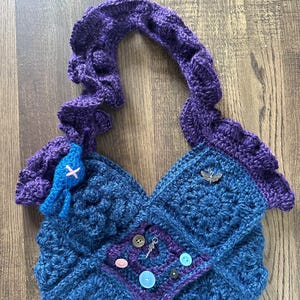 Op de afbeelding: Handgemaakte gehaakte tas in blauwe en paarse tinten. De tas heeft een uniek ontwerp met een textuurpatroon, decoratieve knopen en een kleine bedel. De handgreep is een dikke, gevlochten stijl. Een kleine blauwe versiering is aan de voorkant bevestigd.