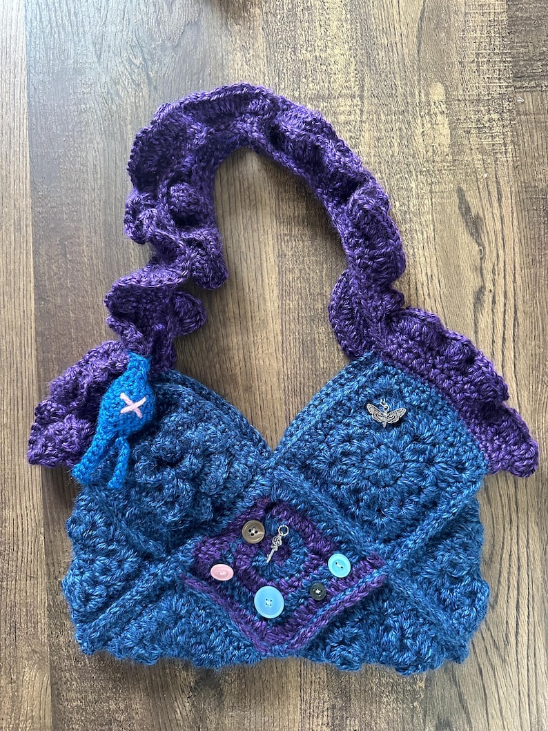 Op de afbeelding: Handgemaakte gehaakte tas in blauwe en paarse tinten. De tas heeft een uniek ontwerp met een textuurpatroon, decoratieve knopen en een kleine bedel. De handgreep is een dikke, gevlochten stijl. Een kleine blauwe versiering is aan de voorkant bevestigd.