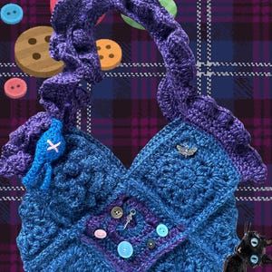 Op de afbeelding: Een handgemaakte blauwe gehaakte tas met een paarse handvat en accenten. De tas heeft een vierkant ontwerp met diverse knopen en een kleine bedel. Een blauw vogelachtig versiering en een zilveren vlinderbedel zijn ook aanwezig.