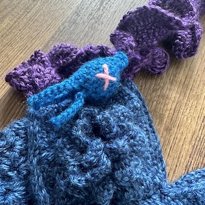 Op de afbeelding: Handgemaakte gehaakte top in blauwe en paarse tinten. De top heeft een blauwe vogelapplicatie met een roze kruis. Het haakwerk heeft een getextureerd uiterlijk en het ontwerp bevat een knoopsluiting. De top is waarschijnlijk een draagbaar kunstwerk.