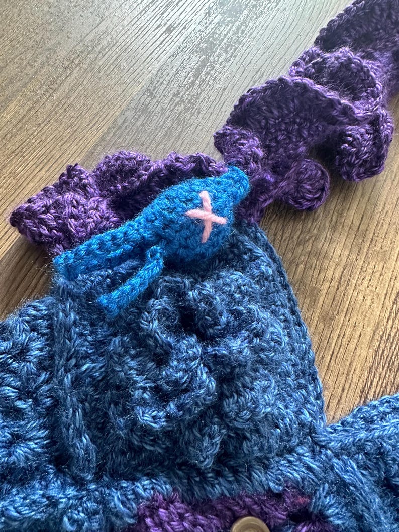Op de afbeelding: Handgemaakte gehaakte top in blauwe en paarse tinten. De top heeft een blauwe vogelapplicatie met een roze kruis. Het haakwerk heeft een getextureerd uiterlijk en het ontwerp bevat een knoopsluiting. De top is waarschijnlijk een draagbaar kunstwerk.