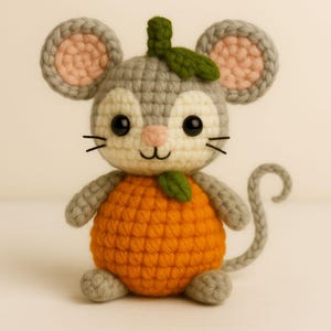May include: A handmade crochet mouse toy with a gray head, ears, and tail, and an orange body. The mouse has black eyes, a pink nose, and a green leaf on its head. The toy is designed to look like an orange.