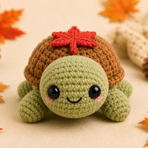 Pode incluir: Uma pelúcia de tartaruga feita em crochê com cabeça e corpo verde-claros, apresentando grandes olhos pretos e bochechas rosadas. A tartaruga tem um casco marrom coberto por uma única folha de bordo vermelha em crochê. O brinquedo é arredondado.