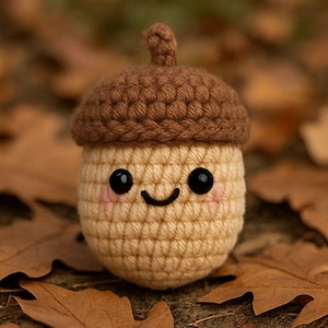 May include: A crocheted acorn with a smiling face, featuring a brown cap and a beige body. The acorn has black eyes and pink cheeks. The background includes fallen brown leaves, creating a fall theme.