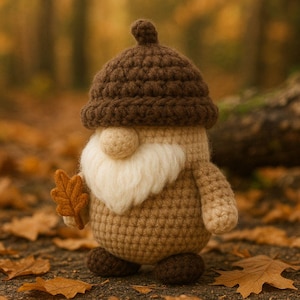 Puede incluir: Una muñeca de gnomo de ganchillo con un sombrero marrón en forma de bellota, una barba color crema y un cuerpo beige. El gnomo sostiene una pequeña hoja naranja. La muñeca está de pie sobre una superficie cubierta de hojas caídas, con un fondo borroso de bosque.