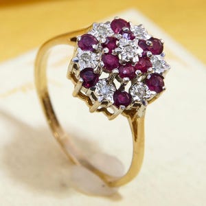 May include: A gold ring with a cluster of gemstones. The ring has a floral design with small, round, deep red rubies and clear diamonds. The band is polished gold. The ring's design is reminiscent of a flower.