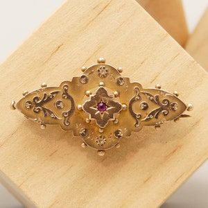 Puede incluir: Un broche de oro con una piedra preciosa roja en el centro y diseños florales intrincados. El broche está engastado con pequeñas piedras redondas.