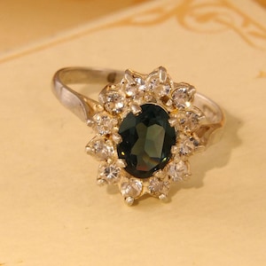 May include: A silver ring with a large oval-shaped dark green gemstone surrounded by a cluster of smaller clear gemstones.