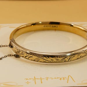 Könnte beinhalten: Ein goldfarbener Klapparmreif mit eingraviertem Blattmuster auf der Außenseite. Das Armband verfügt über eine dünne Sicherheitskette am Verschlussmechanismus und zeigt den Text CMR auf der Innenseite des Bandes.