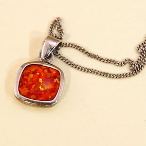 Könnte beinhalten: Eine silberne Halskette mit einem quadratischen Anhänger mit einem leuchtend orangefarbenen Edelstein. Der Anhänger ist in eine silberne Fassung eingefasst und hängt an einer zarten Silberkette. Der Edelstein hat ein funkelndes, strukturiertes Aussehen.