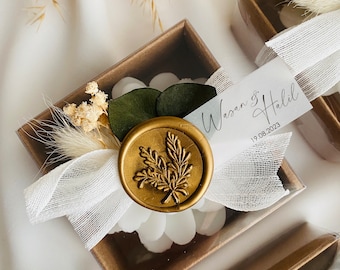 Könnte beinhalten: Eine quadratische braune Geschenkbox mit einer weißen blütenförmigen Kerze, verziert mit einem weißen Band, Trockenblumen, einem dunkelgrünen Blatt und einem goldenen Wachssiegel mit Blattmotiv. Ein weißes Etikett trägt die Aufschrift Wasan & Halil 19.08.2023.