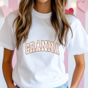 May include: White t-shirt featuring the word "Granna" in a pink, floral, and brown design. The text is in a collegiate style font. The shirt has a crew neck and short sleeves.