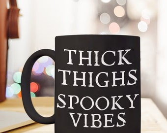 May include: A black ceramic mug featuring the white text "THICK THIGHS SPOOKY VIBES" printed in a stylised, jagged font on the front. The mug has a standard cylindrical shape with a curved handle on the left side, displaying a matte finish.