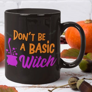 May include: Black ceramic mug with the text "Don't Be A Basic Witch" in orange and purple. The mug is surrounded by orange pumpkins, acorns, and fall leaves, creating a seasonal theme.