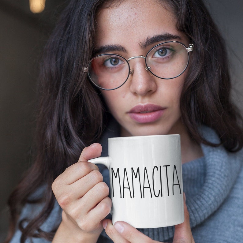 May include: White ceramic mug with the word "MAMACITA" in black, handwritten-style lettering. The mug has a curved handle and is held by a person wearing a gray sweater. The mug is a standard size for coffee or tea.