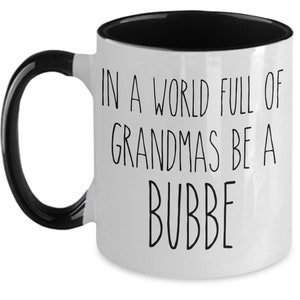 May include: White ceramic coffee mug with a black handle and black rim. The mug has the text "In a world full of grandmas be a bubbe" printed on it.