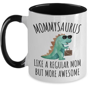 May include: White ceramic coffee mug with a black rim and handle. The mug has a green dinosaur with sunglasses and a purse printed on it. The text "Mommyosaurus like a regular mom but more awesome" is printed on the mug.