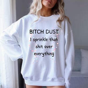 Puede incluir: Sudadera blanca con el texto "BITCH DUST I sprinkle that shit over everything" en negro. El texto está rodeado por un borde decorativo de estrellas en colores pastel. La sudadera tiene mangas largas y cuello redondo.