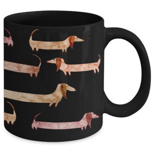 May include: Black ceramic mug featuring a repeating pattern of watercolor dachshunds in shades of brown, tan, and pink. The dogs are depicted with long bodies and floppy ears against a solid black background. The mug has a curved handle.