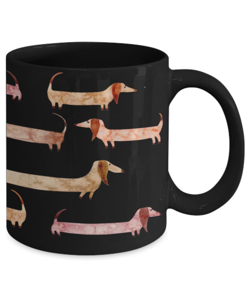 May include: Black ceramic mug featuring a repeating pattern of watercolor dachshunds in shades of brown, tan, and pink. The dogs are depicted with long bodies and floppy ears against a solid black background. The mug has a curved handle.