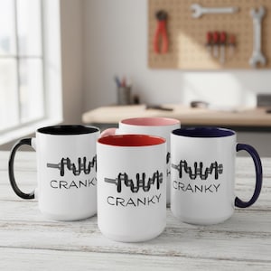 May include: Four white ceramic mugs with different coloured interiors and handles. Each mug features a black graphic of an engine crankshaft and the word "CRANKY" in black text. The mug interiors are black, pink, red, and blue.