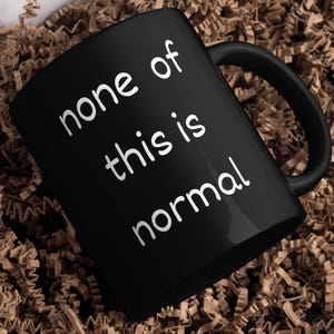 May include: Black ceramic mug with the words "none of this is normal" in white, sans-serif font. The mug has a curved handle and sits on a bed of brown paper shreds. A simple, yet bold design.