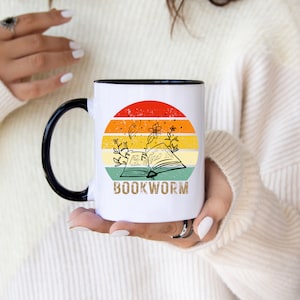 May include: White ceramic mug with a black rim and handle. The mug features a retro-style sunset design with a book and flowers. The text "BOOKWORM" is printed in brown on the mug.