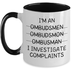 May include: A white ceramic mug with a black interior and handle. The front features black text that reads: I'M AN OMBUDSMEN (crossed out), OMBUDSMON (crossed out), OMBUSMAN (crossed out), I INVESTIGATE COMPLAINTS. The design is printed in a simple font.