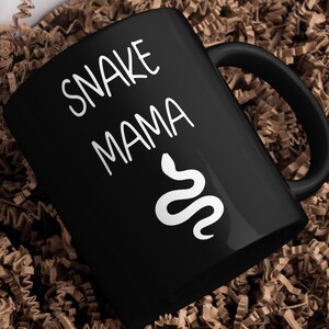 May include: Black ceramic mug featuring the text "SNAKE MAMA" in white, accompanied by a white snake design. The mug has a black handle and is placed on a bed of brown paper. Ideal for reptile enthusiasts.