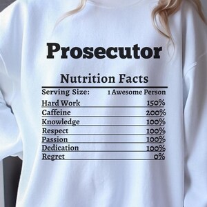 May include: White sweatshirt with the word "Prosecutor" in bold black letters. Below, the text "Nutrition Facts" is followed by a list detailing the composition of an "Awesome Person," including Hard Work, Caffeine, and Dedication.