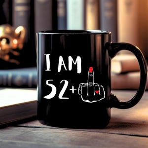 Puede incluir: Taza de café negra con texto blanco que dice "I AM 52+" y un dedo medio blanco con un corazón rojo en la uña.