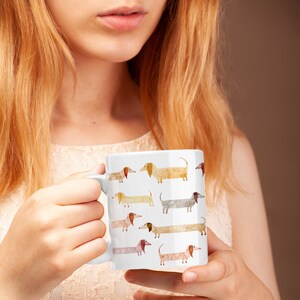 May include: White ceramic mug with a colorful watercolor design of dachshunds. The dachshunds are in various poses and colors, including brown, tan, and gray.