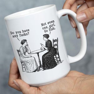 May include: A white ceramic mug featuring a black and white vintage-style illustration of two women sitting at a table. The text reads: Do you have any fucks? and Not even one, girl. Go fish. The mug has a curved handle and a smooth, glossy finish.