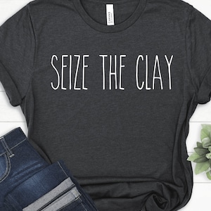 以下が含まれることがあります： 前面に白い文字で「Seize the Clay」とプリントされた濃いグレーのTシャツです。