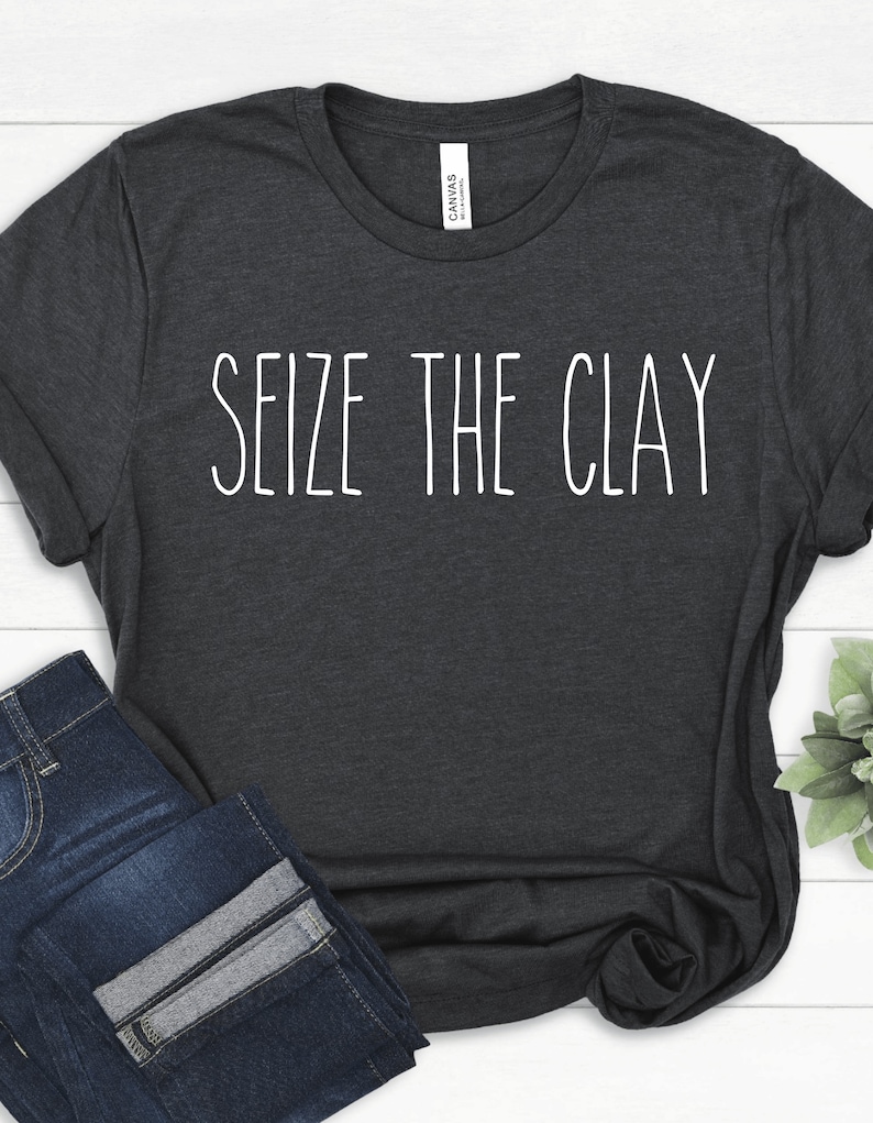 以下が含まれることがあります： 前面に白い文字で「Seize the Clay」とプリントされた濃いグレーのTシャツです。