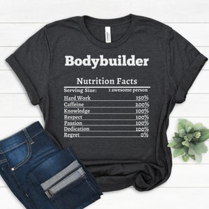 Peut inclure: T-shirt gris foncé avec le mot "Bodybuilder" en blanc. En dessous, une étiquette "Nutrition Facts" liste des ingrédients comme "Hard Work" à 150% et "Regret" à 0%. Un jean bleu plié est visible.