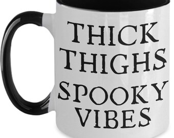 May include: White ceramic mug with a black interior, handle, and rim. The mug features the text "THICK THIGHS SPOOKY VIBES" in bold black letters. The mug is designed for hot beverages.